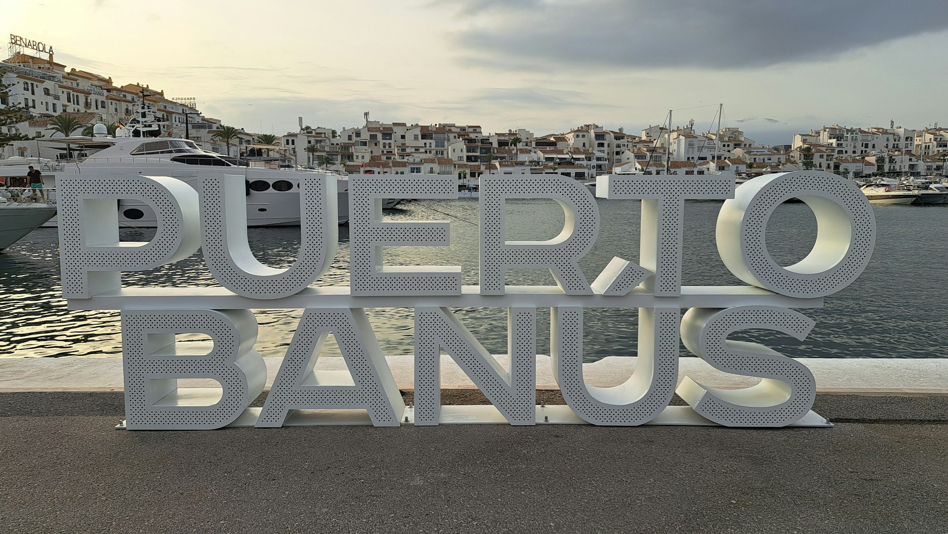 A metal sign that says puerto banus in front of a body of water